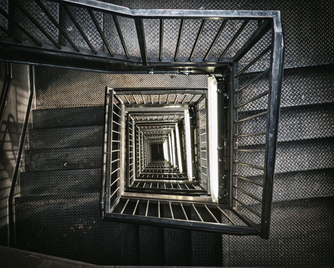 A view from the top, looking down into an industrial building stairwell. Metal stairs and railings lead down several floors into the basement, representing the legal situation of a second staircase