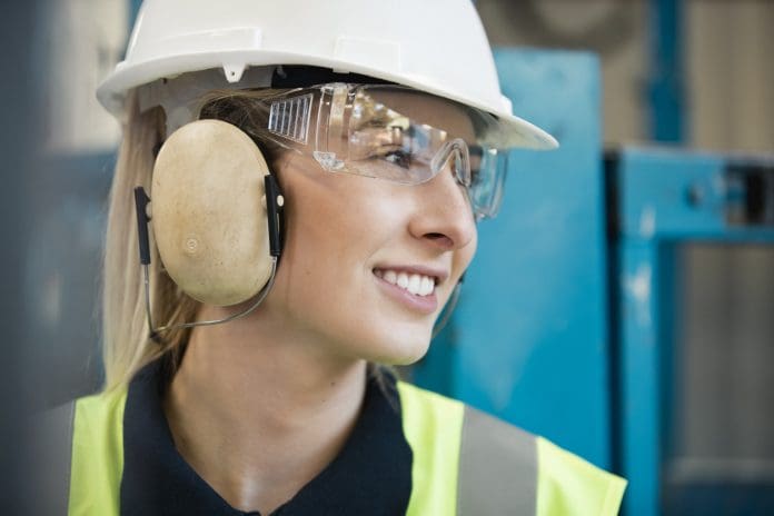 A close-up shot of a young woman at work, she is wearing a hard hat, protective ear muffs, protective eyeglasses​ and a hi-vis jacket.