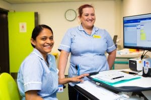 Two smiling nurses in light blue uniforms work at a computer station in a hospital, with one nurse seated and the other standing beside her