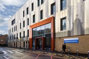 Exterior of the Willowfield Building at King’s College Hospital, featuring a modern facade with beige and metallic panels, a prominent red-framed glass entrance, and a blue NHS sign on the wall.