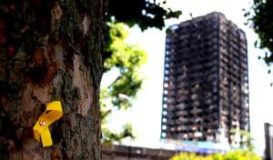 Grenfell Tower, a high rise building severely damaged by fire, in the background with a Yellow Ribbon commemorating those lost pinned to a tree