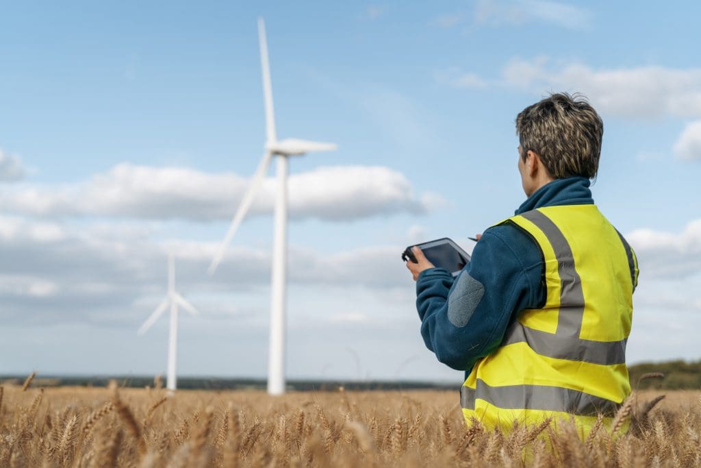 A technician in a high-visibility vest observes wind turbines from a wheat field, monitoring data on a tablet.