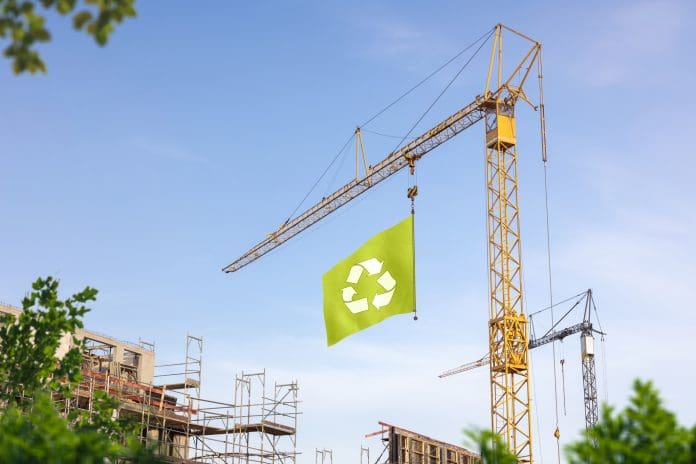 A construction site with a crane hoisting a green flag with a recycling symbol.