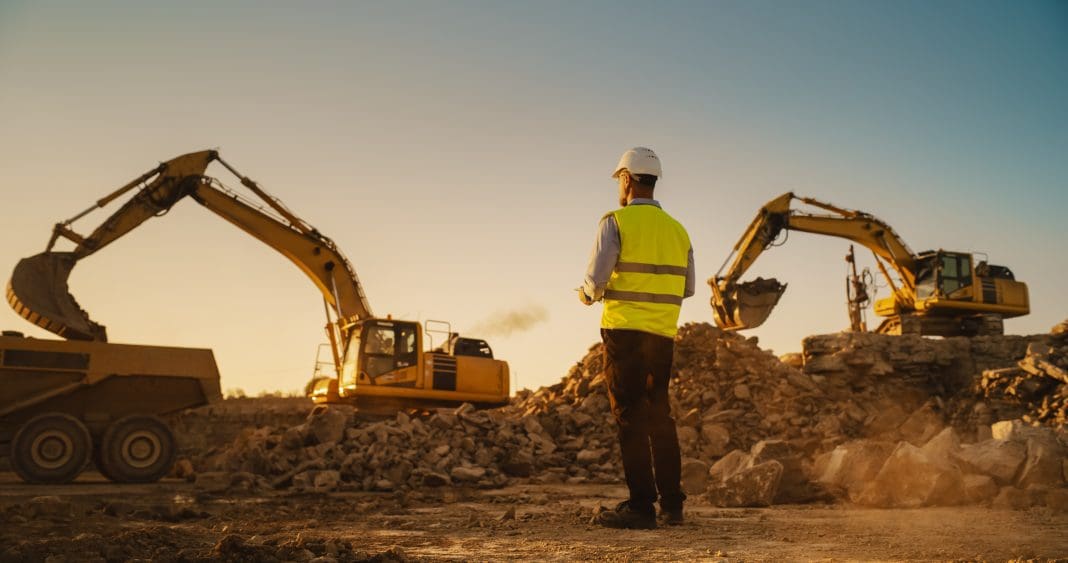 Caucasian Male Urban Planner Wearing Protective Goggles And Using Tablet On Construction Site On A Sunny Day. Man Inspecting Building Progress. Excavator Loading Materials Into Industrial Truck, representing construction administrations