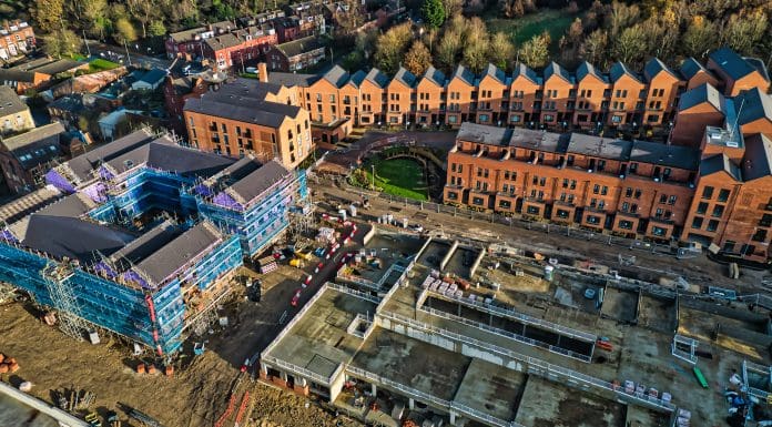 Aerial view of a residential construction site. Brick townhouses are complete, while adjacent buildings are under construction, surrounded by scaffolding and building materials. The scene shows a mix of finished and unfinished structures, representing topics to be discussed at BEYOND '25 LIVE.