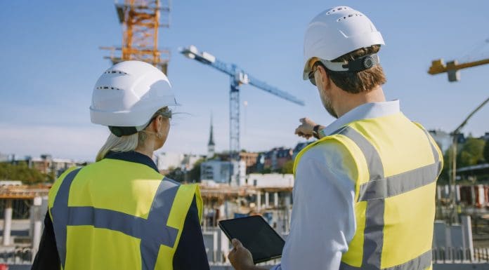 Male Civil Engineer and Young Female Building Architect Use a Tablet Computer on a City Construction Site. They Talk About the Future of Real Estate Development and Planning. Wearing Safety Hard Hats.