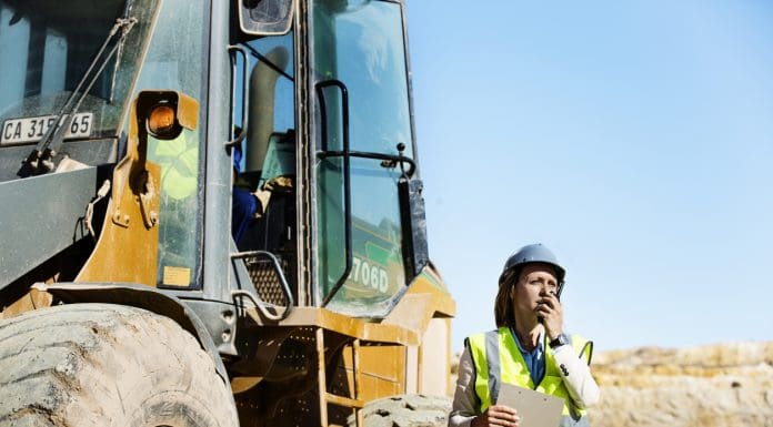 Female architect using walkie-talkie by bulldozer at quarry against clear sky, representing fleet insurance costs