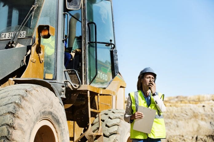 Female architect using walkie-talkie by bulldozer at quarry against clear sky, representing fleet insurance costs