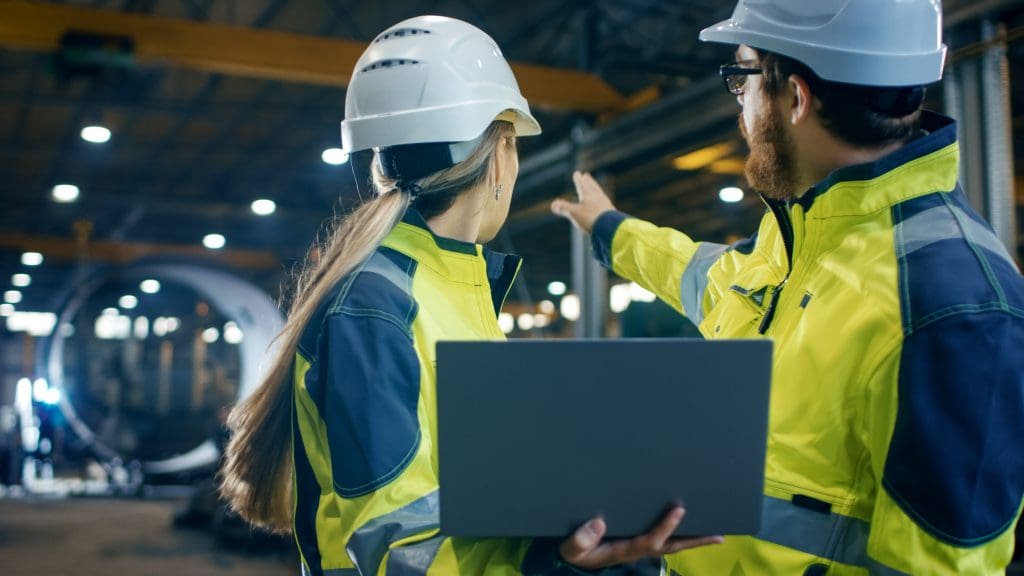 Inside the Heavy Industrial Factory Female Industrial Engineer Holds Laptop and Has Discussion with Project Manager. They Wear Hard Hats and Safety Jackets. In the Background Welding/ Metalwork in Progress.