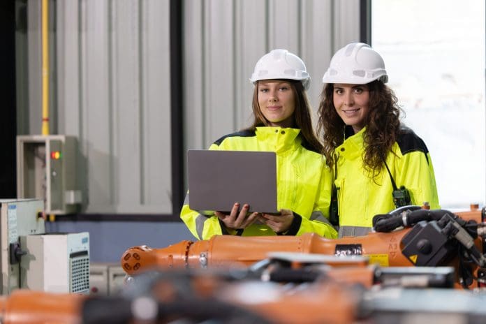 Two engineers in safety gear analyze robotic arms in a modern manufacturing plant. They check configurations and ensure quality standards are met, representing professional indemnity insurance